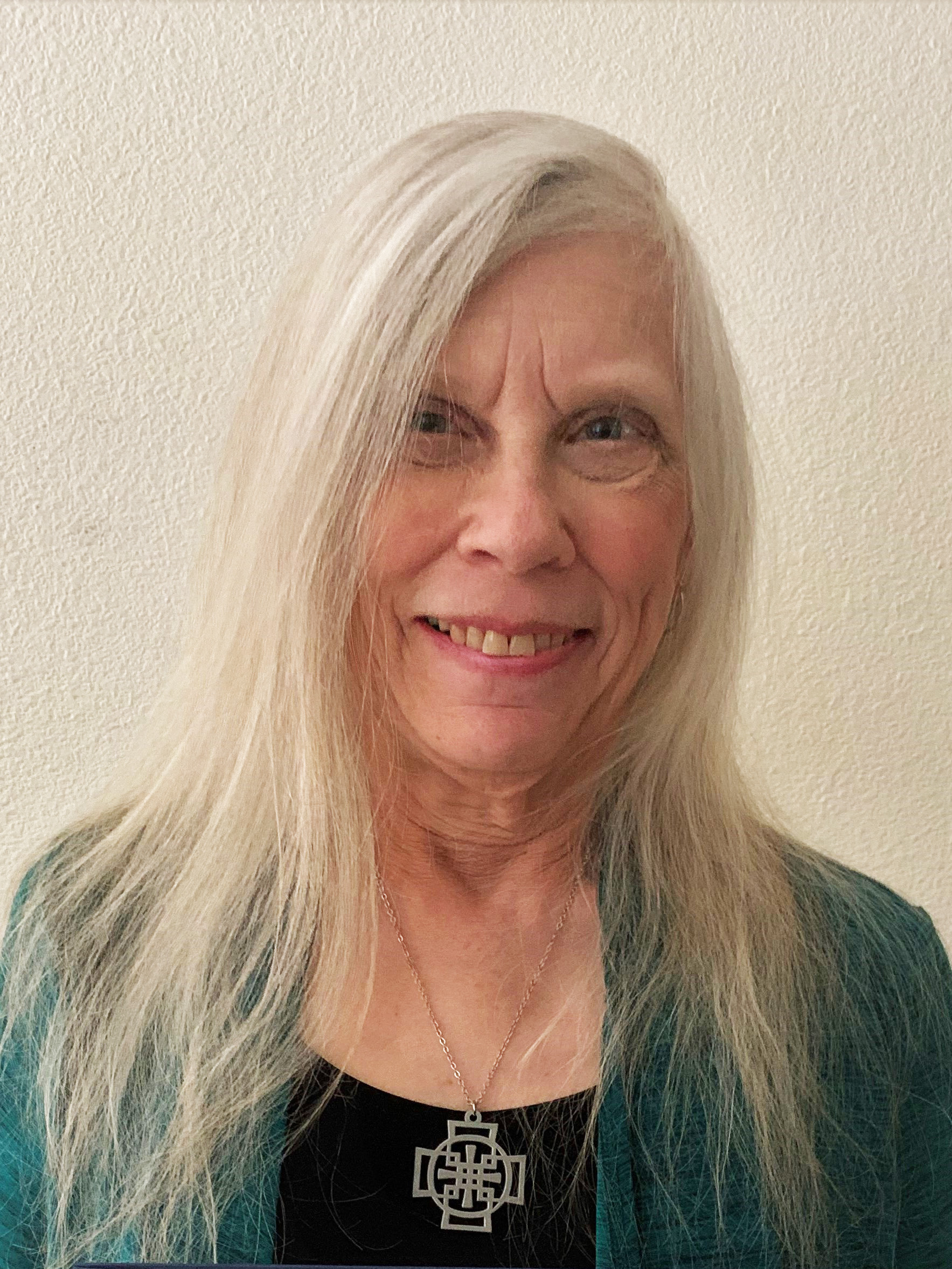 An holder women with long straight white hair smiles for the camera wearing a green sweater and black shirt with a cross pendant on. 