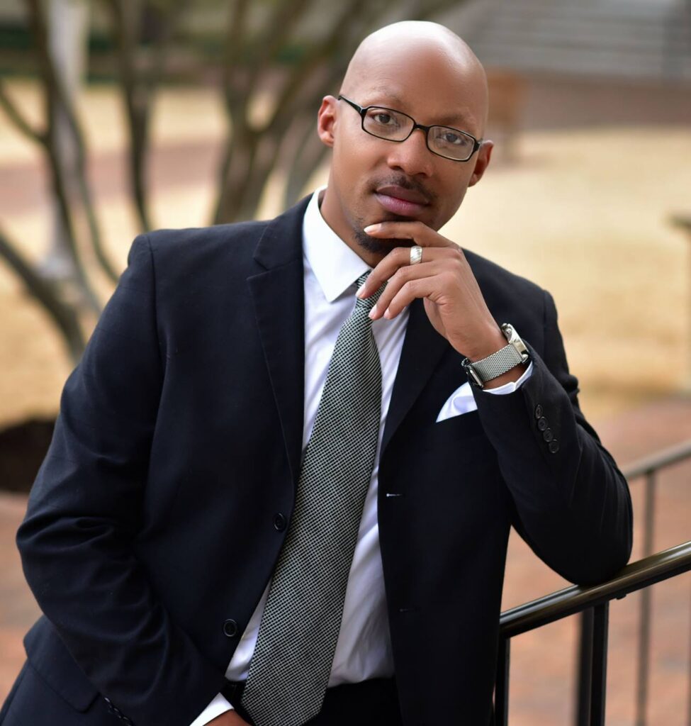 An african american man with shaved head, goatee, and glasses, wearing a suit, stands with his hand under his chin on a stairway.