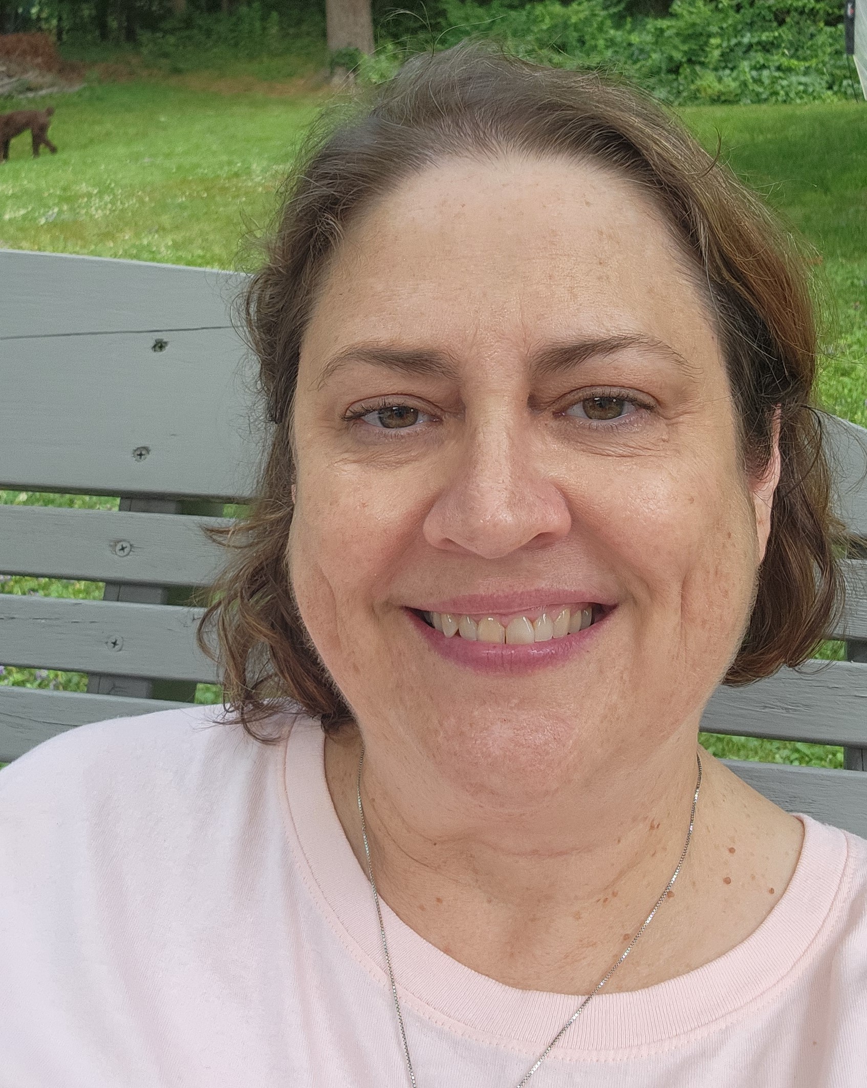 An older woman with brown shoulder length hair wearing a pink shirt smiles for the camera.
