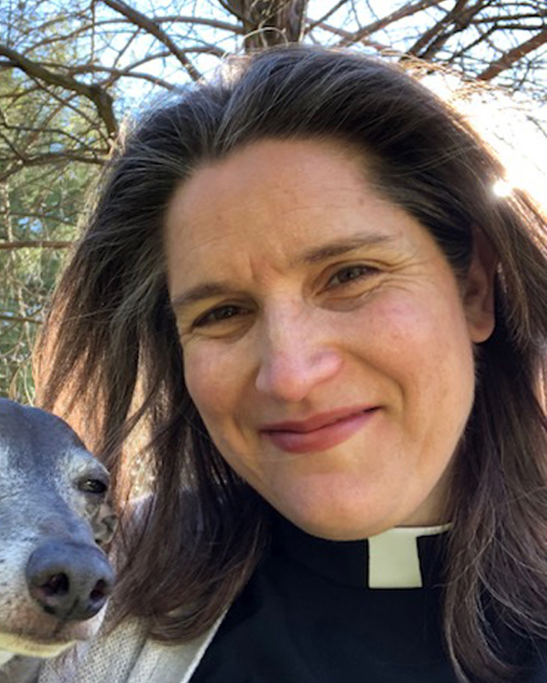 A women with shoulder length brown hair wearing a pastoral collar smiles for the camera with a small dog to her left. 