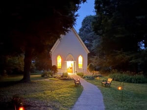 A small white chapel in the woods on a dark evening with its windows lit up.